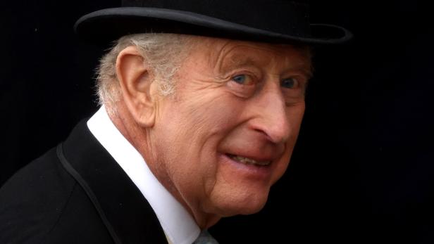 König Charles III. trägt einen schwarzen Hut und einen Anzug vor einem dunklen Hintergrund.