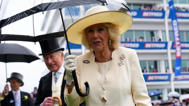 Königin Camilla hält bei Regen einen Regenschirm, während König Charles III. im Hintergrund bei einem Pferderennen steht.