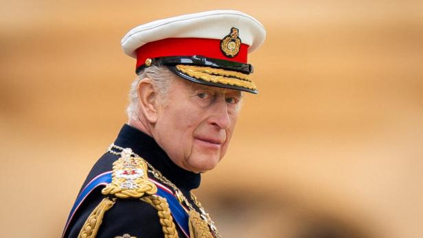 König Charles III. trägt eine militärische Uniform mit einer weißen Schirmmütze und goldenen Abzeichen vor einem hellen Hintergrund.