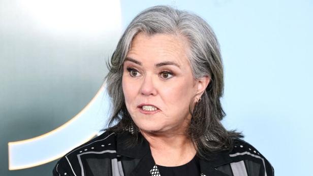 Rosie O’Donnell mit grauem Haar trägt einen gemusterten Blazer und zeigt mit dem Finger nach vorne.