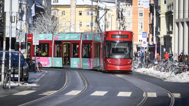 Eine rot-türkise Straßenbahn fährt durch eine winterliche Stadtstraße mit vielen geparkten Fahrrädern und Fußgängern am Rand.