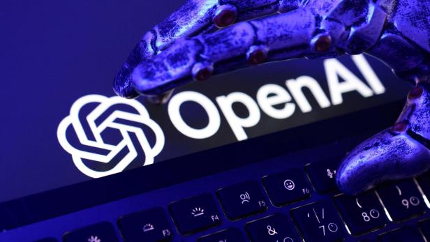 Eine metallische Roboterhand schwebt über einer Tastatur, im Hintergrund ist das OpenAI-Logo zu sehen.