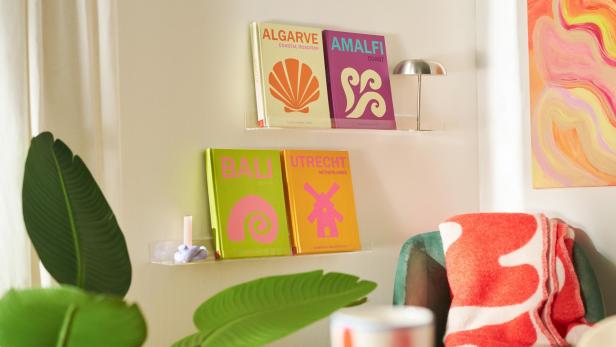 Vier farbenfrohe Fotobücher mit den Titeln Algarve, Amalfi, Bali und Utrecht stehen dekorativ auf Wandregalen in einem gemütlich eingerichteten Wohnzimmer.