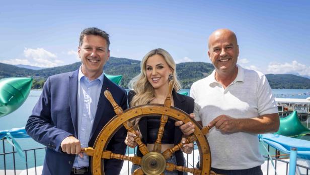 Drei Personen halten gemeinsam ein großes hölzernes Steuerrad am Wörthersee.