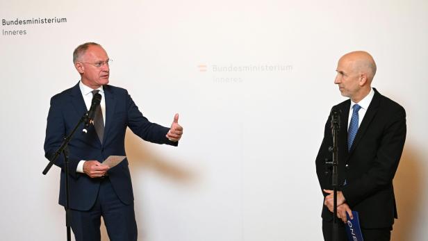 Zwei Männer im Anzug stehen vor Mikrofonen und sprechen miteinander vor einer weißen Wand mit Behördenaufschrift.