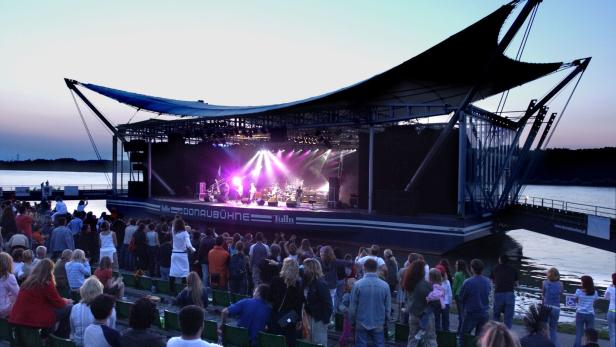 Viele Menschen verfolgen ein Open-Air-Konzert auf einer modernen Bühne am Wasser bei Sonnenuntergang.