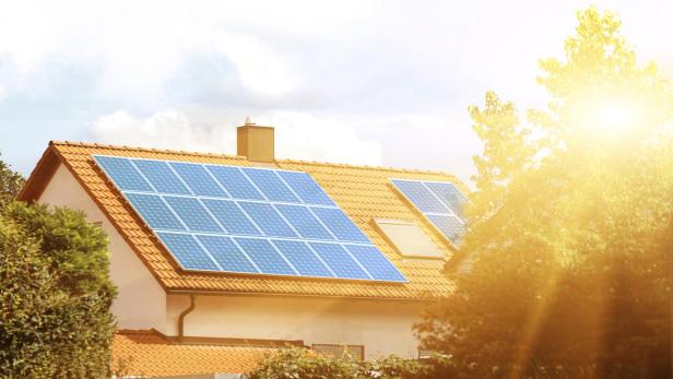 Auf einem Hausdach mit roten Ziegeln sind mehrere Solarpanels installiert, Sonnenstrahlen scheinen darauf.