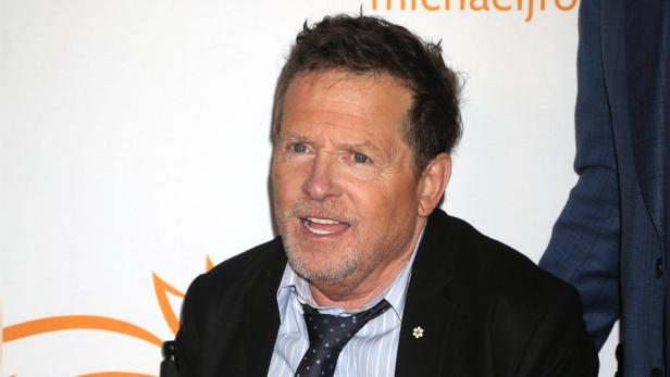 Michael J. Fox trägt ein hellblau gestreiftes Hemd mit Krawatte und ein schwarzes Sakko vor einem hellen Hintergrund.