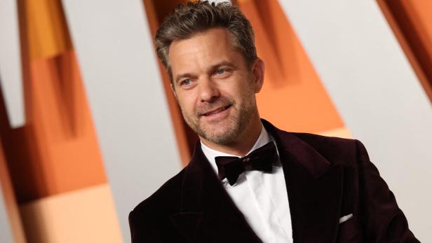 Joshua Jackson trägt einen dunkelroten Samt-Smoking mit Fliege vor einem orangefarbenen Hintergrund.