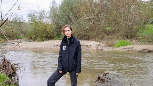 Eine Person steht am Ufer eines Flusses vor dichtem, herbstlichem Gebüsch und trägt eine schwarze Jacke mit WWF-Logo.