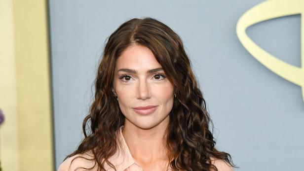 Janet Montgomery lächelt in pfirsichfarbener Bluse vor hellblauem Hintergund.