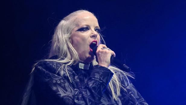 Shirley Manson singt mit hellem Haar und auffälligem Augen-Make-up in ein Mikrofon vor einem dunklen, blauen Hintergrund.