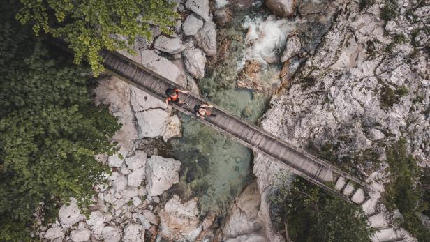 Luftaufnahme einer schmalen Holzbrücke über einen felsigen Bach mit zwei darauf sitzenden Personen und umgebendem Grün.