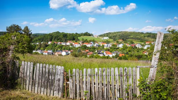 Ein altes Holzzaun steht vor einer Wiese, dahinter ein Dorf mit roten Dächern und grünen Hügeln unter blauem Himmel.