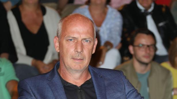 Mario Basler sitzt mit verschränkten Beinen in einem Sessel, im Hintergrund sind mehrere Zuschauer zu sehen.