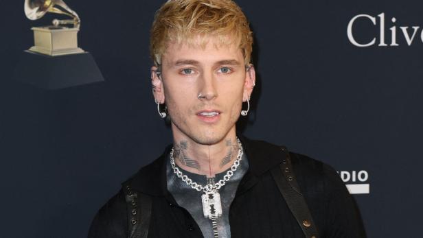 Machine Gun Kelly mit blonden Haaren, Tattoos und auffälliger Halskette vor einem dunklen Hintergrund mit Grammy-Logo.