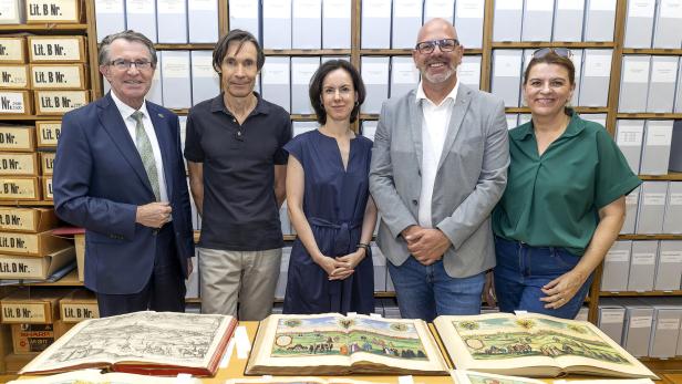 Fünf Personen stehen vor Regalen mit Archivboxen, auf dem Tisch liegen mehrere aufgeschlagene historische Bücher mit Stadtansichten.