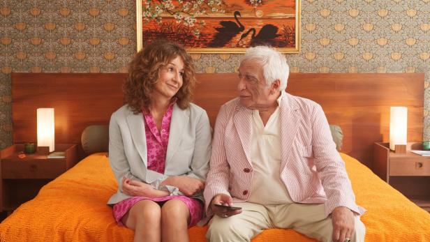 Gérard Darmon und Valérie Lemercier sitzen auf einem Bett und sehen sich an.