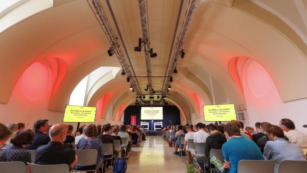Viele Menschen sitzen in einem modernen Saal mit gewölbter Decke und hören einem Vortrag auf einer Bühne mit großen Bildschirmen zu.