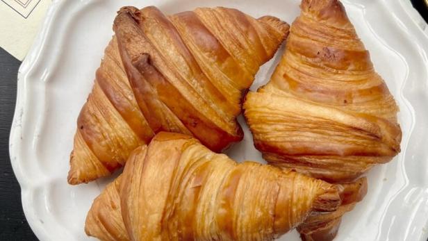 Drei goldbraune, knusprige Croissants liegen auf einem weißen, gewellten Teller vor einem dunklen Hintergrund.