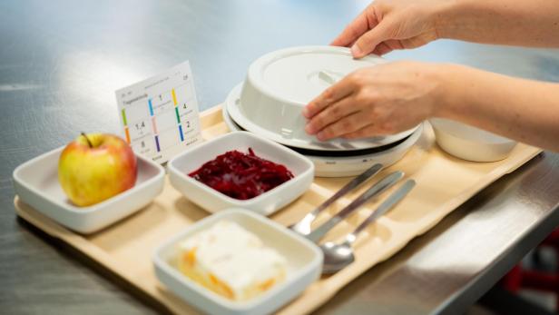 Ein Tablett mit Apfel, Rote-Bete-Salat, Dessert, Besteck und einer abgedeckten Schale wird von einer Person vorbereitet.