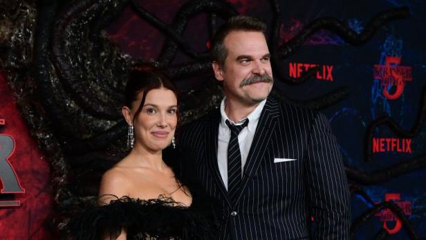 Millie Bobby Brown und David Harbour posieren vor einem Stranger Things-Hintergrund von Netflix.