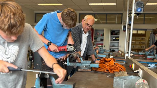 Zwei junge Burschen arbeiten mit Metall in einer Werkstatt und werden dabei von einem Lehrer beaufsichtigt.