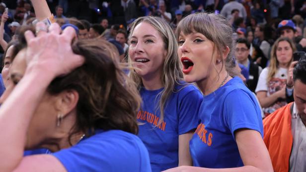 Taylor Swift und eine Begleiterin jubeln in blauen T-Shirts bei einem Basketballspiel in einer vollen Arena.