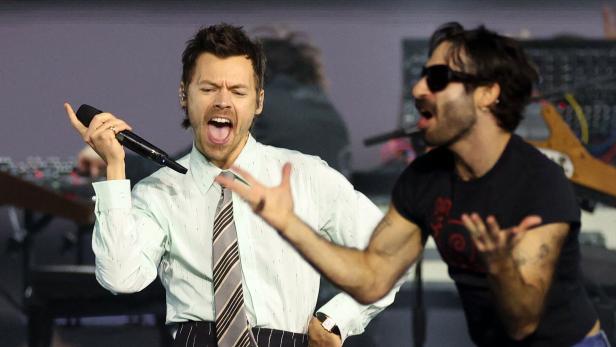 Harry Styles singt mit einem Mikrofon auf einer Bühne, während ein Mann mit Sonnenbrille neben ihm gestikuliert.