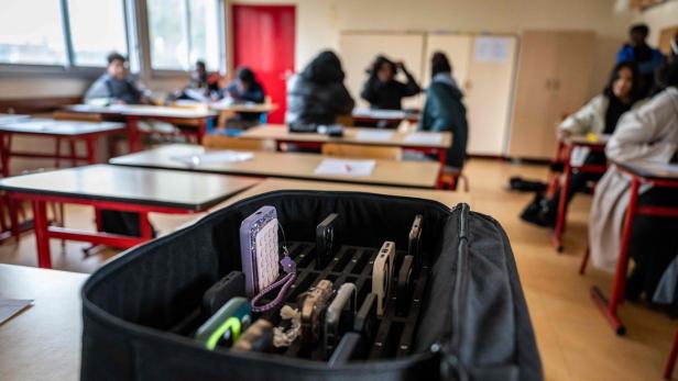 Mehrere Smartphones stecken in einer Aufbewahrungstasche, während Schüler an Tischen im Hintergrund sitzen.