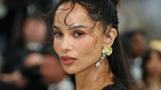 Zoë Kravitz trägt ein schwarzes Spitzenoberteil und auffällige goldene Ohrringe vor einem unscharfen Hintergrund.