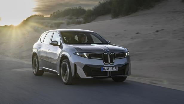 Ein silberner BMW Neue Klasse iX3 fährt auf einer Straße an einer Dünenlandschaft vorbei.