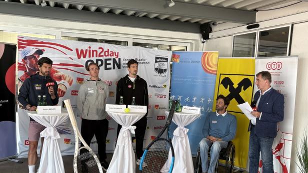Vier Männer stehen und einer sitzt im Rollstuhl bei einer Pressekonferenz mit Tennisbezug vor Sponsorenwänden und großen Tennisschlägern.