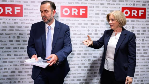 Zwei Anzugträger stehen vor einer ORF-Wand, der Mann hält Unterlagen, die Frau zeigt mit der Hand nach rechts.