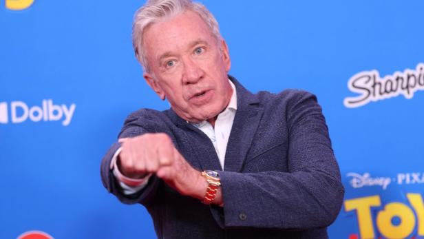 Tim Allen posiert vor einer blauen Wand mit Disney-Pixar-Logos und streckt seine Faust in die Kamera.