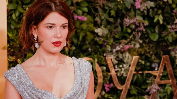 Millie Bobby Brown trägt ein silbernes Kleid vor einer mit Pflanzen und goldenen Buchstaben verzierten Wand.