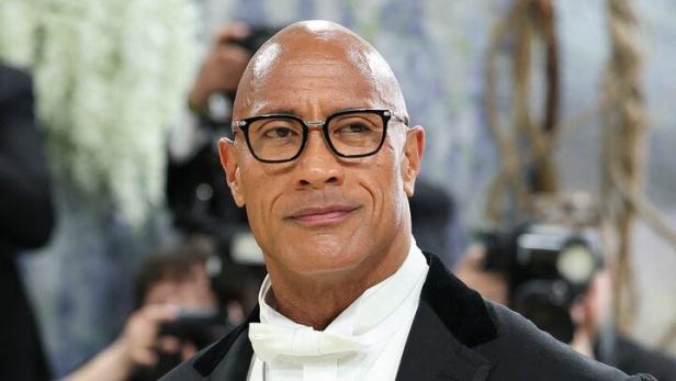 Dwayne Johnson trägt einen eleganten schwarzen Smoking mit weißer Weste, Fliege, Brille und einer auffälligen Brosche vor unscharfem Hintergrund.