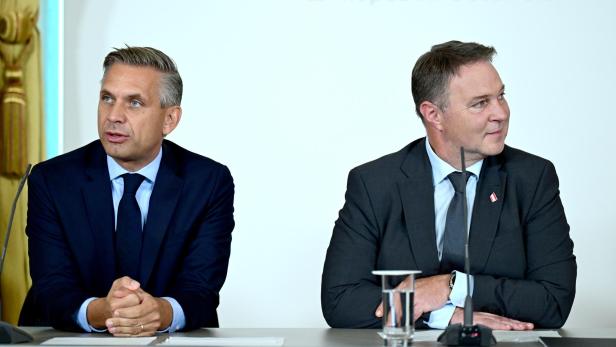 Zwei Männer im Anzug sitzen an einem Tisch mit Mikrofonen und einem Glas Wasser vor neutralem Hintergrund.