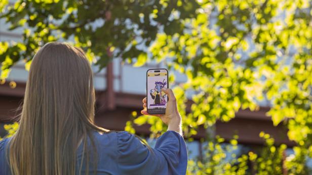 Eine Person mit langen Haaren hält ein Smartphone hoch, auf dessen Bildschirm eine bunte Comicfigur zu sehen ist, umgeben von grünen Bäumen.