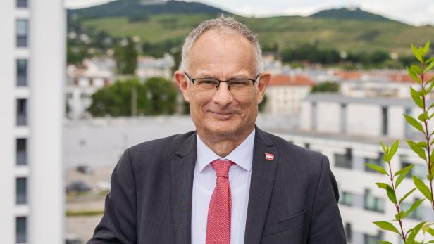 Markus Marterbauer im Anzug mit roter Krawatte steht auf einer Terrasse, im Hintergrund sind Gebäude und grüne Hügel zu sehen.