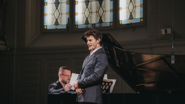 Ein Mann im Anzug singt neben einem Flügel, während ein Pianist spielt, im Hintergrund bunte Kirchenfenster.