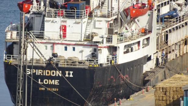 Ein Frachtschiff mit dem Namen SPIRIDON II liegt am Hafen, daneben sind gestapelte Ballen zu sehen.