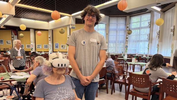 Ein junger Mann vom Verein T.I.W. steht hinter einer Seniorin, die eine VR-Brille trägt, in einem hellen Gemeinschaftsraum.