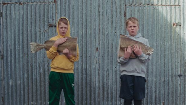 Zwei isländische Buben halten jeweils einen Fisch und schauen in die Kamera.
