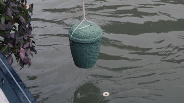 Eine Person hält eine grüne Urne über das Wasser, während Blumen auf der Wasseroberfläche treiben.