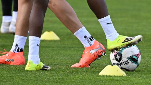 Zwei Fußballspieler mit bunten Schuhen stehen auf dem Rasen, einer hält einen Ball zwischen gelben Markierungshütchen.