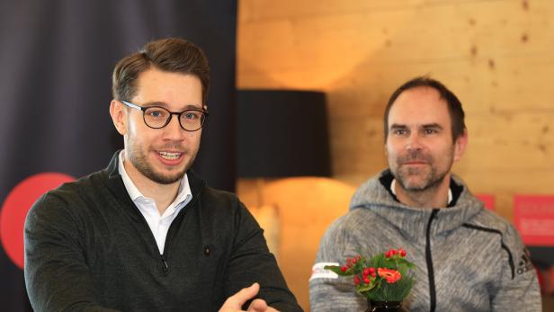 Zwei Männer sitzen an einem Tisch, einer trägt Brille und spricht, vor ihnen steht eine kleine schwarze Vase mit Blumen.