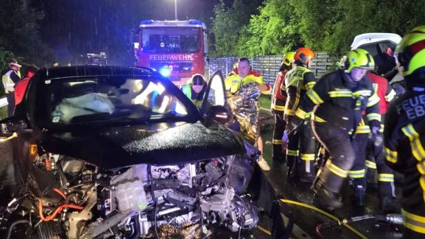 Ein schwer beschädigtes Auto bei Nacht, umgeben von Feuerwehrleuten und Rettungskräften im Einsatz.