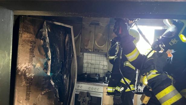 Zwei Feuerwehrleute in Schutzkleidung löschen einen Brand in einer stark beschädigten Küche mit verbrannten Geräten und Möbeln.