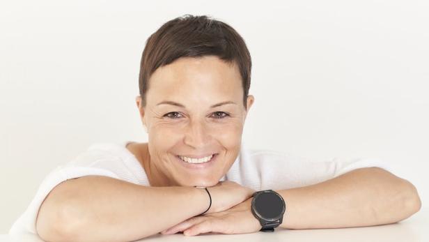 Frau mit kurzen braunen Haaren lächelt und stützt ihr Kinn auf verschränkte Arme, trägt eine schwarze Armbanduhr.